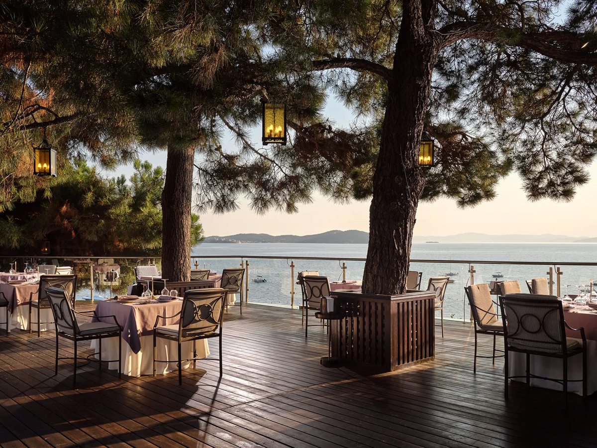 Alfresco restaurant terrace of laid tables on a wooden deck under pine branches, looking out over the calm sea.
