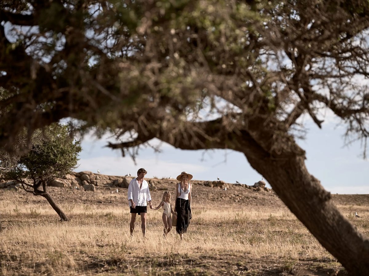 A family walking through an olive grove of dry golden grass and gnarled trees in the resort grounds.
