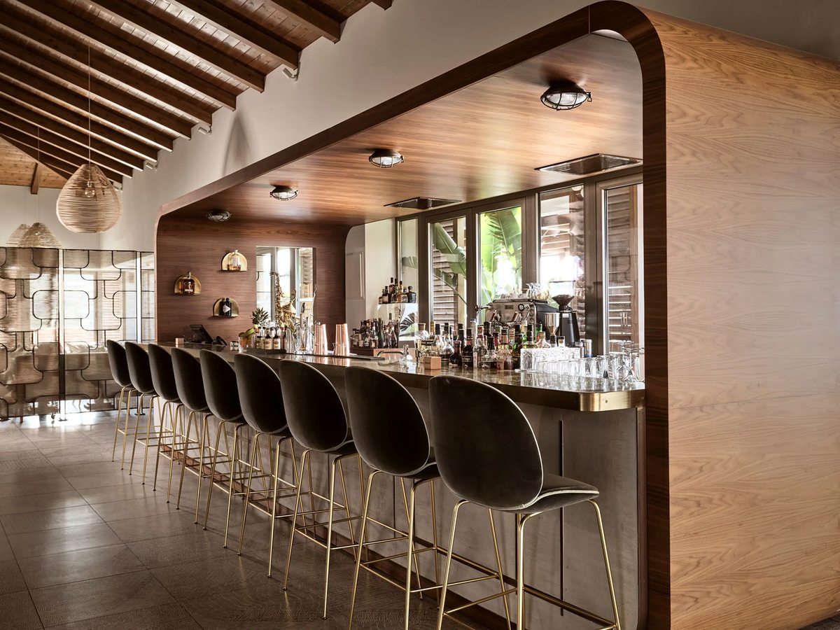 Long indoor cocktail bar with a timber-panelled back wall, spirits and glassware on display and black cushioned stools with brass foot rails.