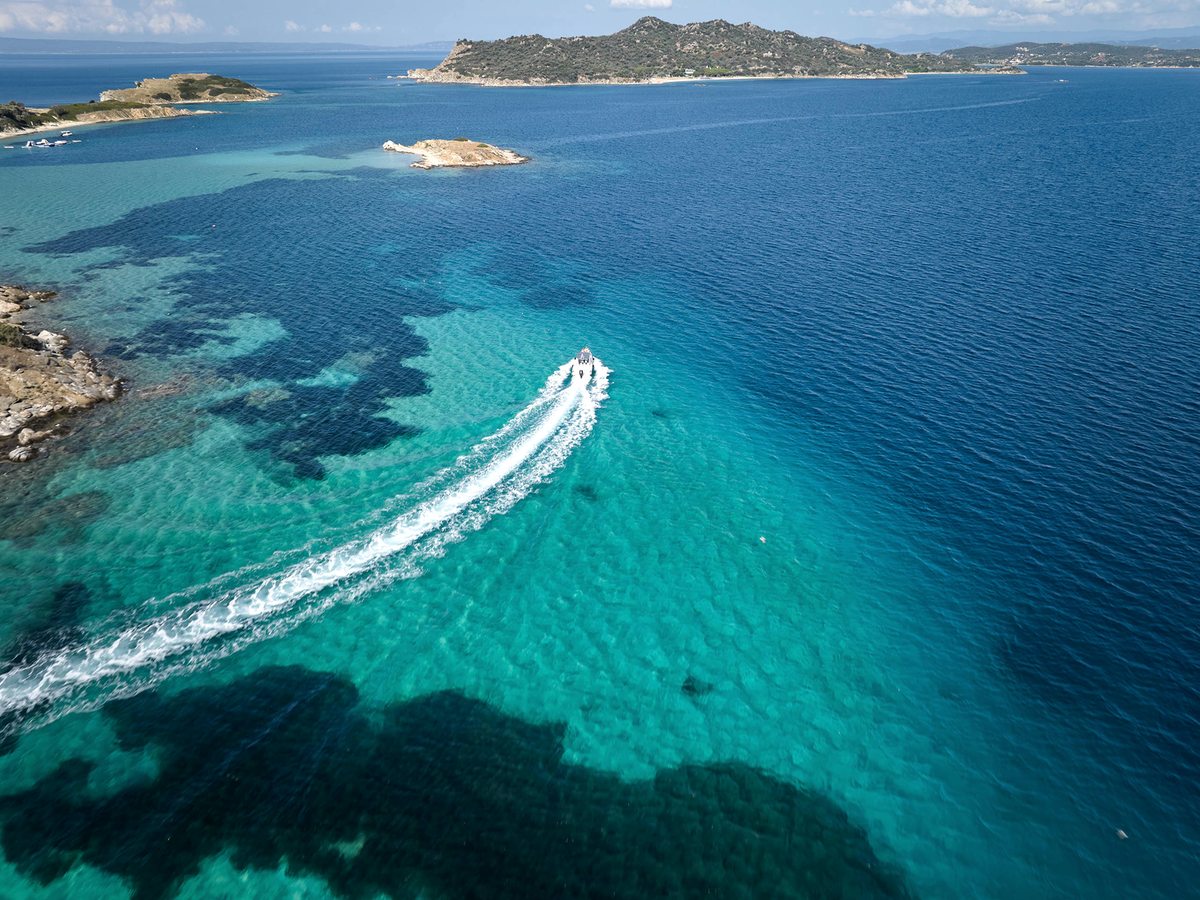 Aerial view of a turquoise bay with a small boat cutting a wake, a rocky shoreline and a green offshore island.
