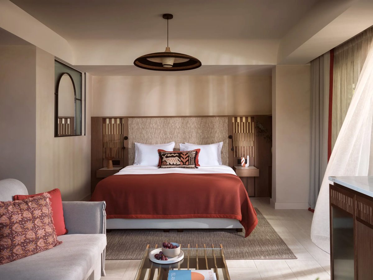 Warm-toned suite bedroom with a double bed, a woven headboard and a rattan pendant light, and a small grey sofa and coffee table in a sitting corner.