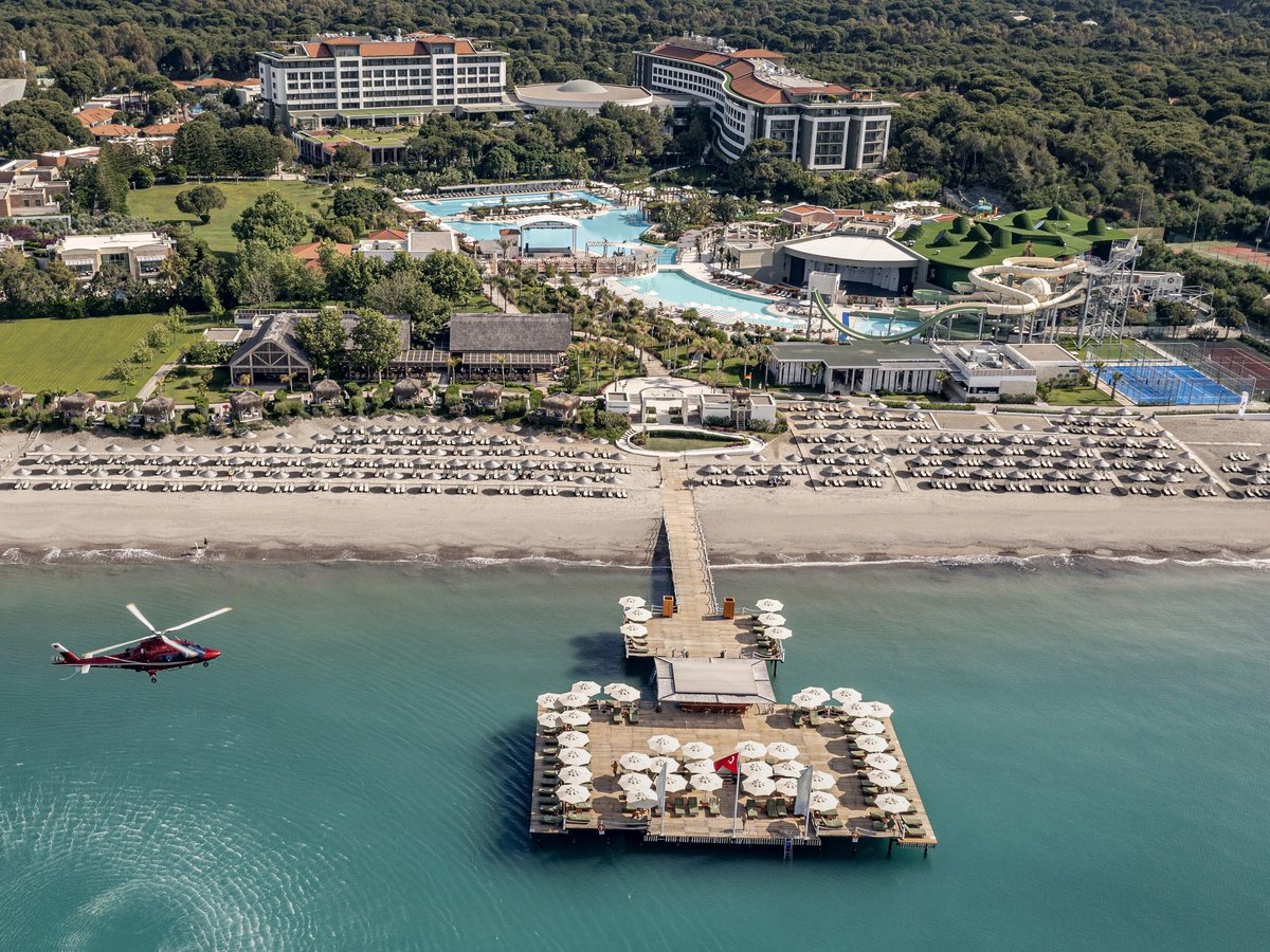 Aerial view of the resort spread along a sandy beach with a pier, pools and low buildings among green grounds, the sea beyond.