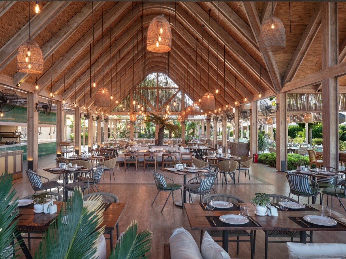 A large open-sided timber pavilion with a high pitched roof, woven pendant lights, a central palm and set tables looking out to the gardens.