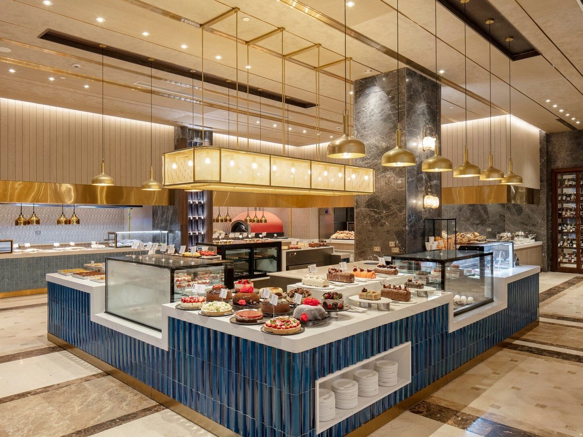 A modern indoor buffet hall with a long blue-fronted serving island of hot dishes and desserts under warm downlights.