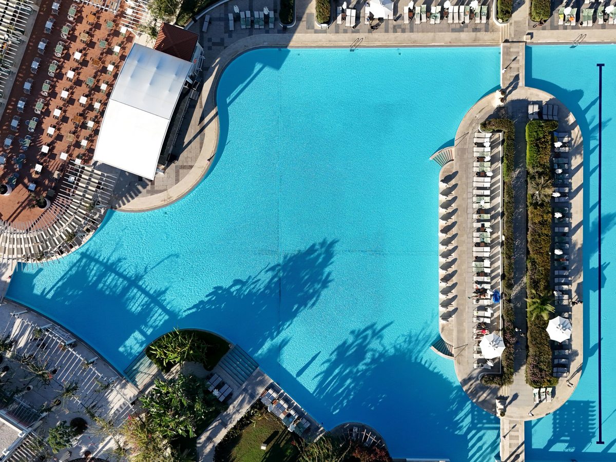 Aerial view of a large blue pool with two long swim-up bar islands, surrounded by decking with the resort behind.