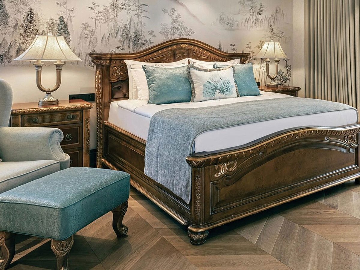 A bedroom with a carved dark-wood king bed, a blue runner and cushions, matching bedside lamps and a small upholstered stool.