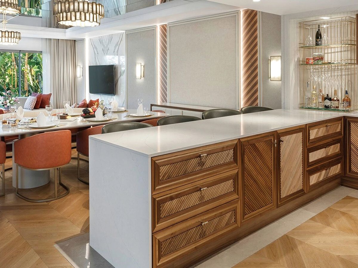 An open-plan suite kitchen with a white island, wooden cabinets, a hob, bar stools and a dining counter beside garden-view windows.