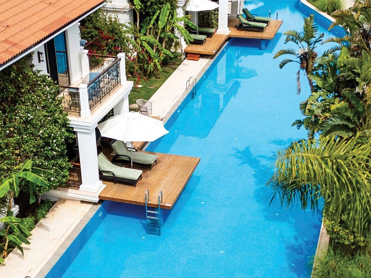 A long narrow blue pool running between balconied buildings, with green sun loungers on wooden decks that step straight into the water.