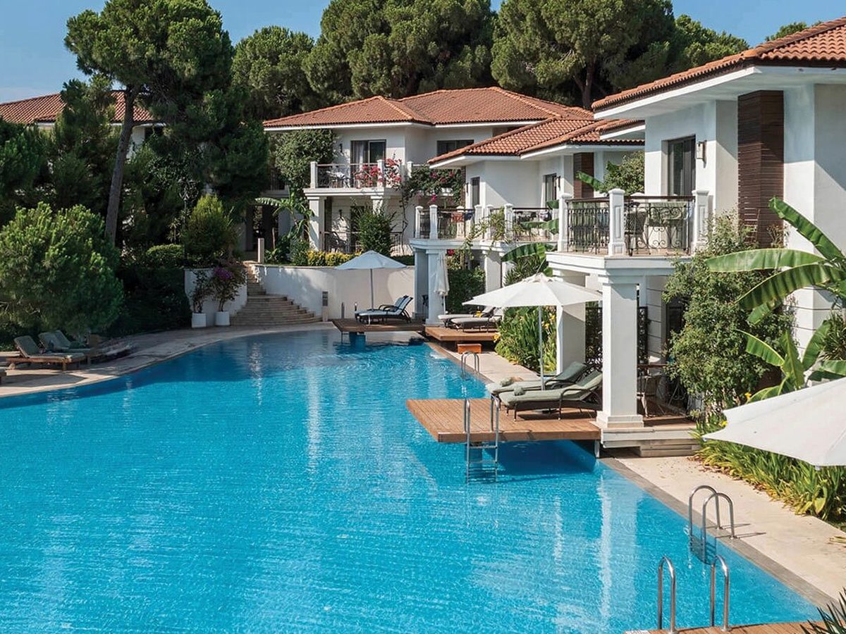 White red-roofed Lake House buildings with balconies around a bright blue pool, with sun loungers on wooden decks among pines and palms.
