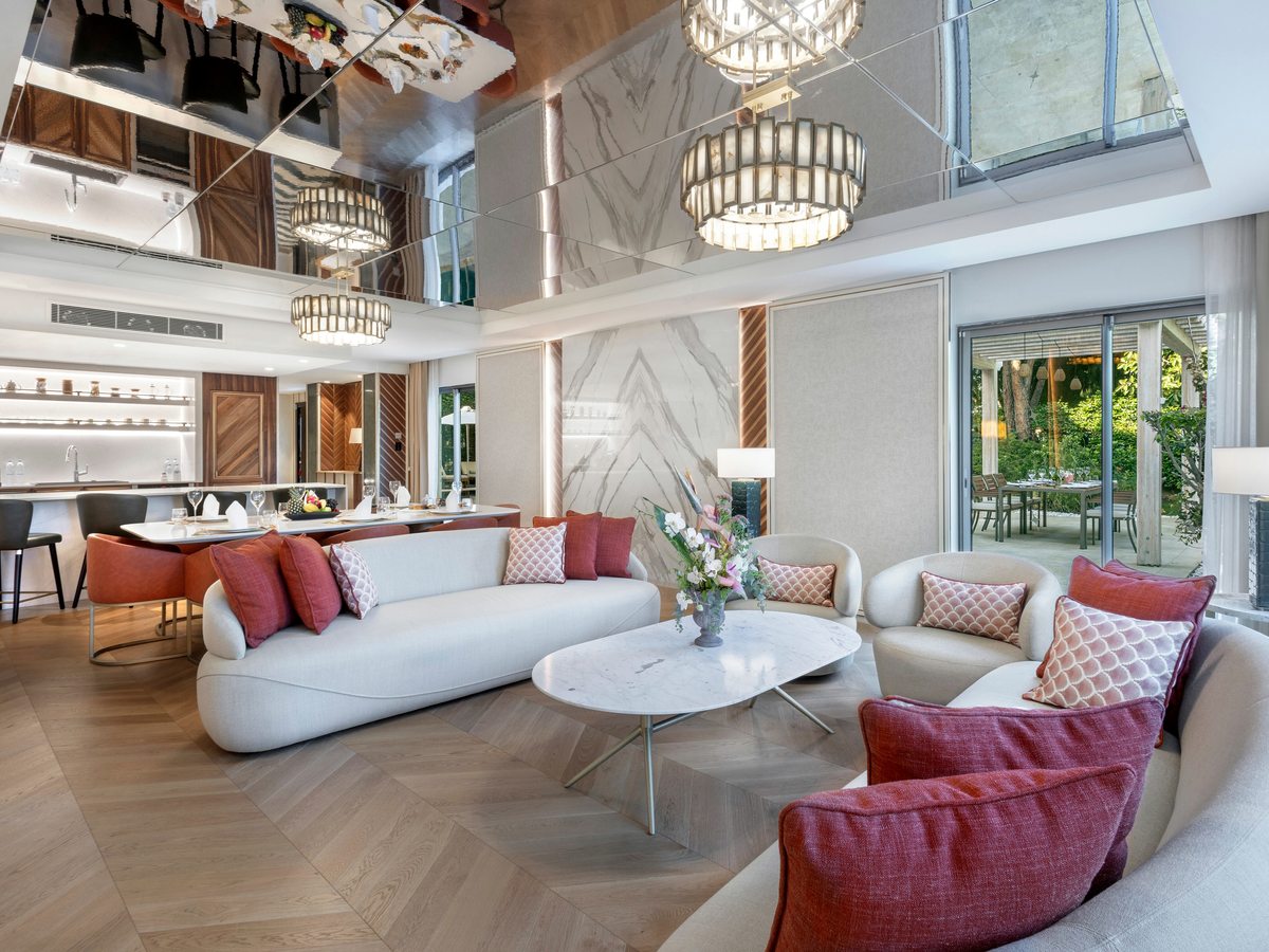 A two-storey suite living room with a mirrored high ceiling, chandeliers, cream sofas with red and pink cushions, a marble coffee table, and an open kitchen and dining area beyond.