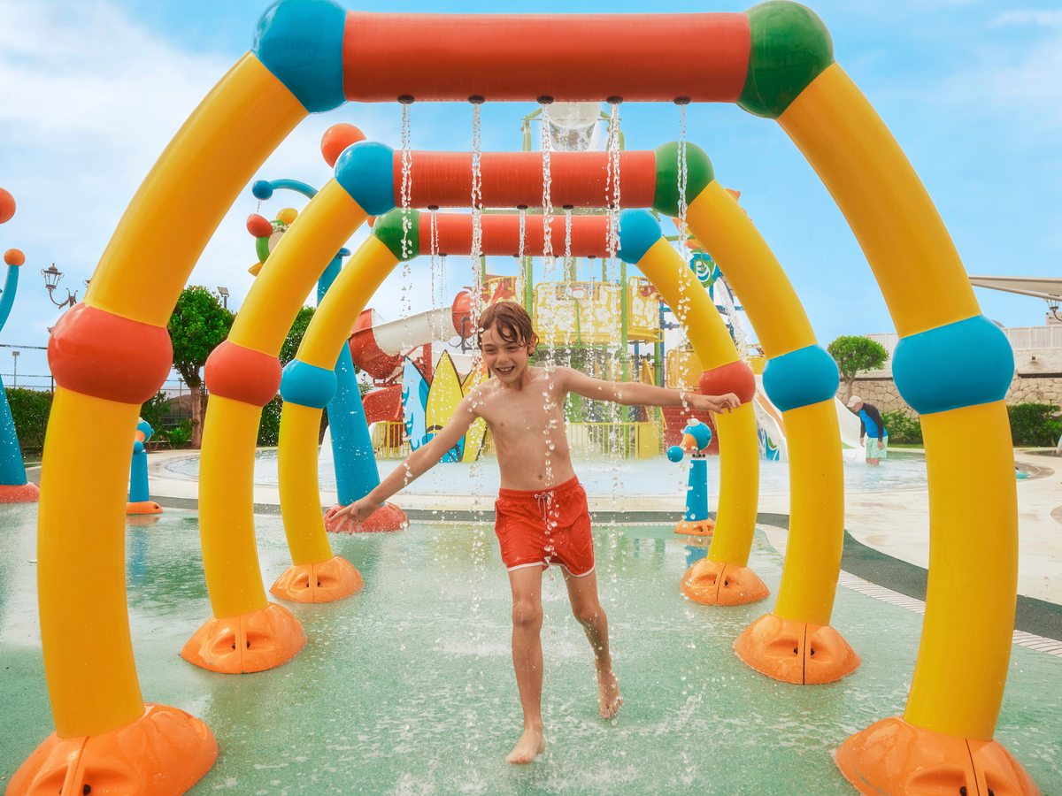 A child in red shorts running through a shallow pool under a colourful inflatable arch of water sprays.