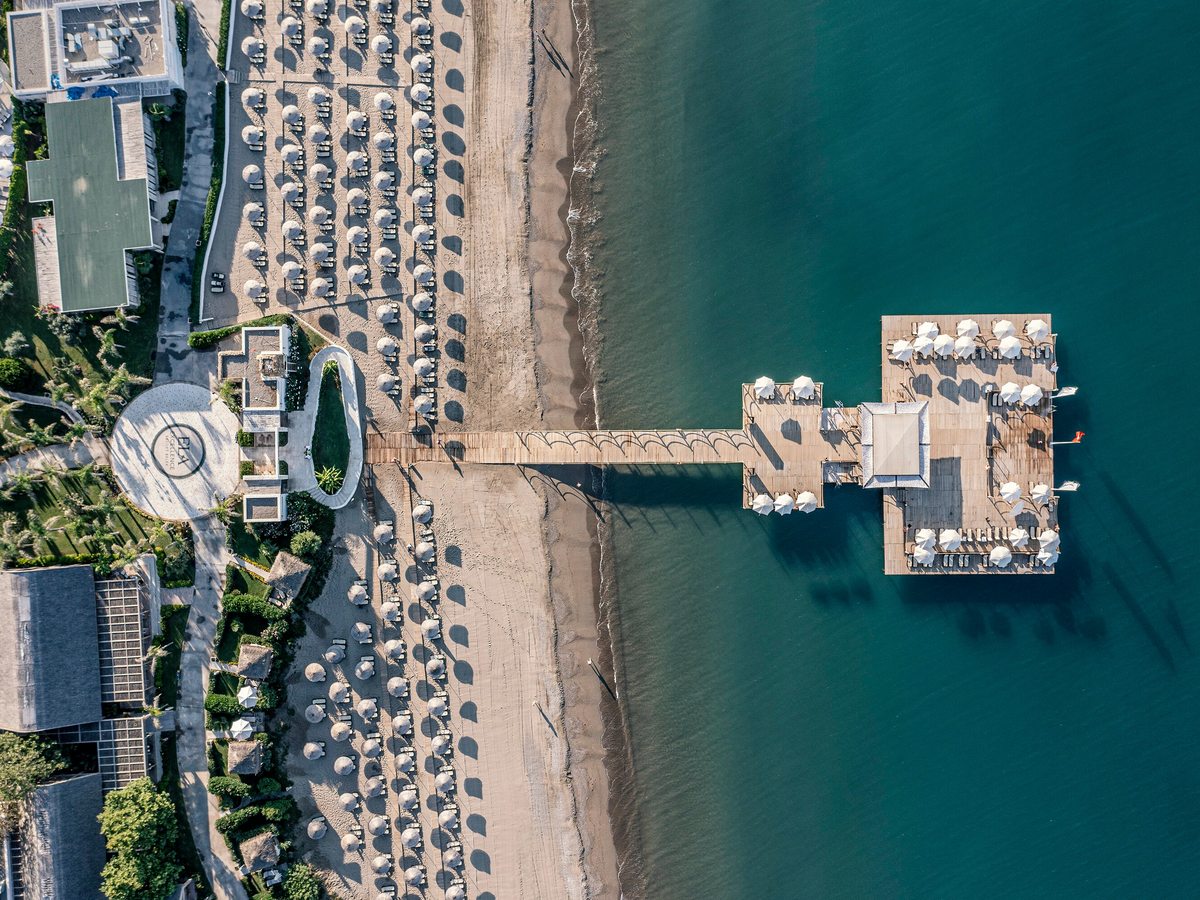 Aerial view of a wooden pier reaching out over turquoise sea to a circular deck, the sandy beach and rows of umbrellas behind.