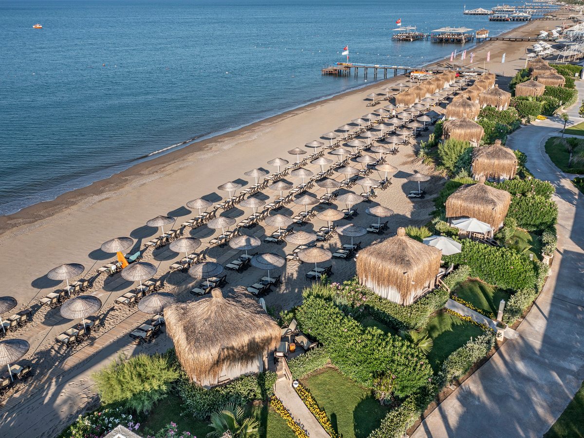 Aerial view of a sandy beach lined with rows of thatched sun umbrellas and cabanas, the calm sea to one side and green grounds to the other.