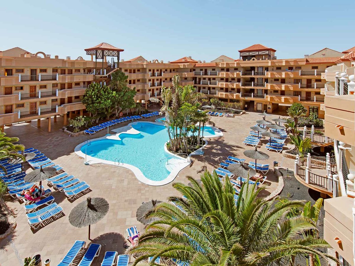 A large freeform swimming pool in the resort courtyard, edged with blue sun loungers and straw parasols and ringed by apartment blocks and palm trees.