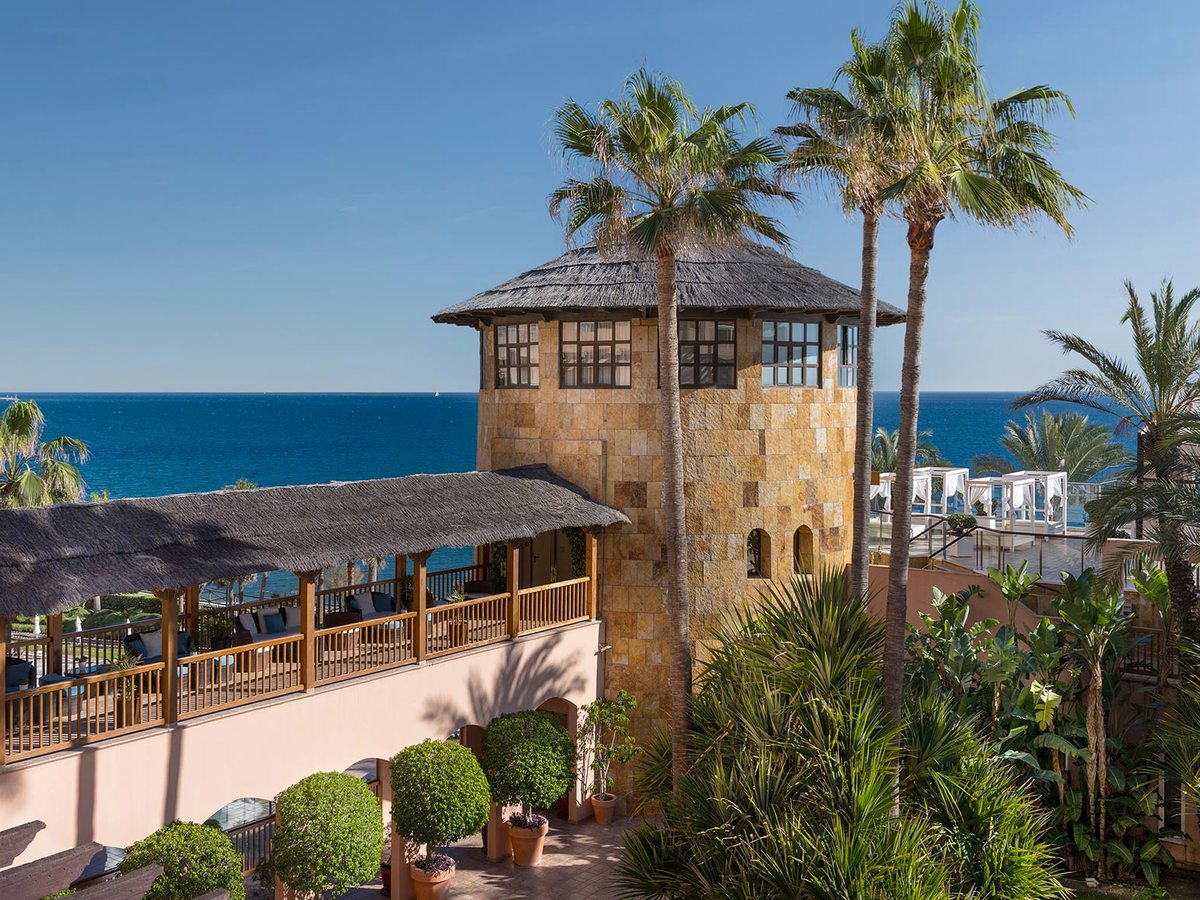 The hotel's stone tower with a thatched-style roof rising above a covered seafront terrace, framed by tall palms under a blue sky with the Mediterranean beyond.