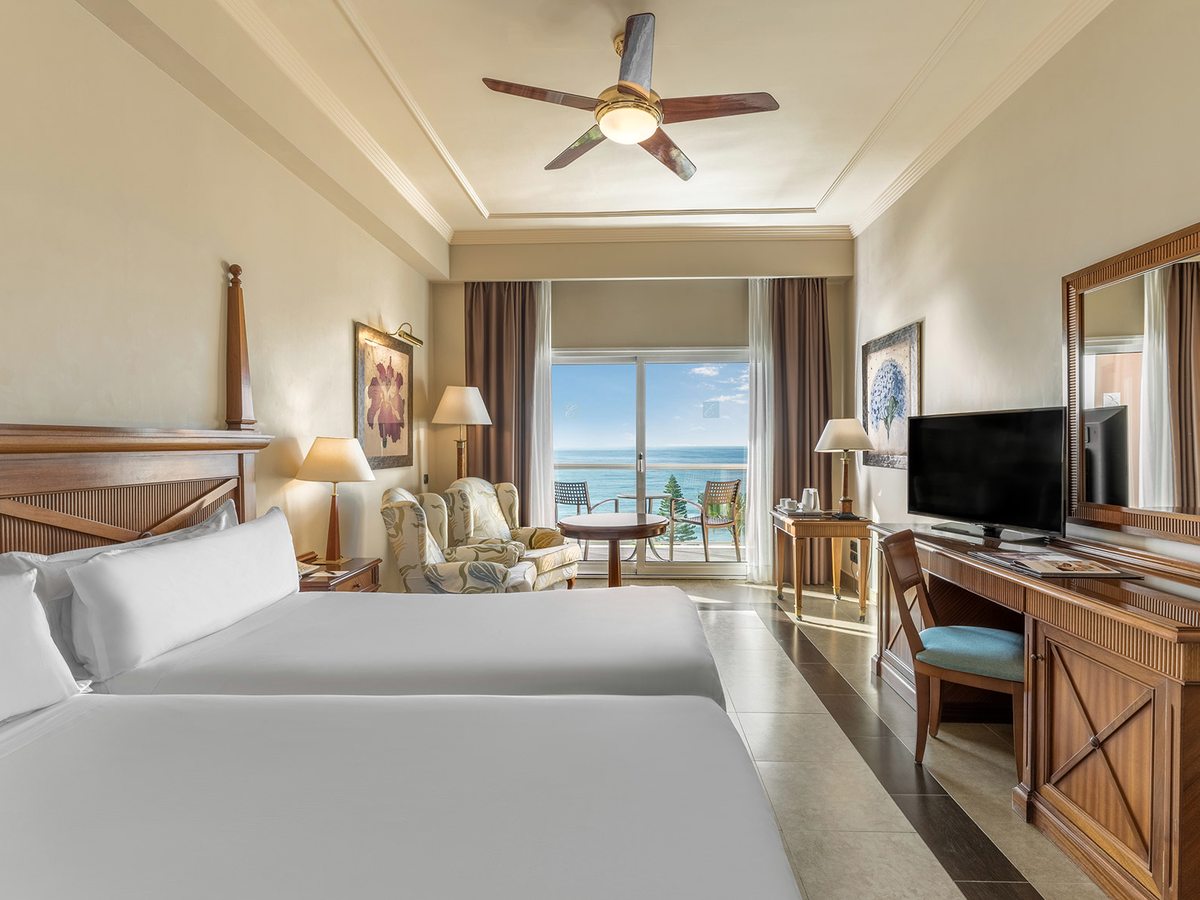 A twin room with two single beds, a sitting area with armchairs, a writing desk, a ceiling fan and a balcony with a sea view.