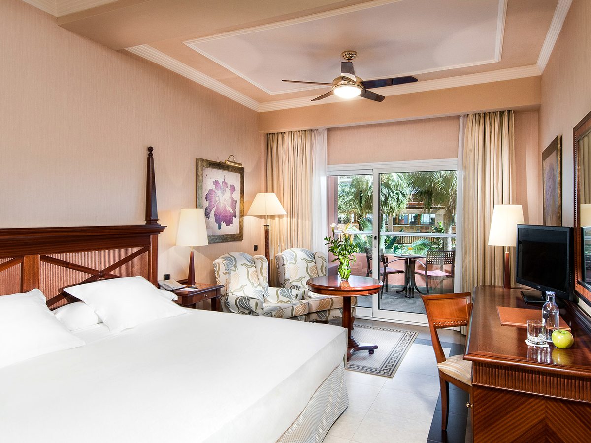 A room with a double bed, warm pink walls, a ceiling fan and sliding doors onto a ground-floor terrace with garden seating.