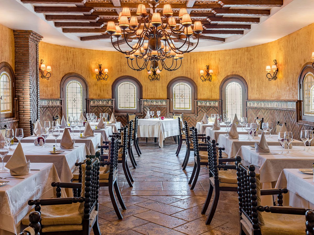 A formal indoor dining room with white-clothed tables, dark wood chairs, arched windows, a chandelier and warm Andalucian-style walls.