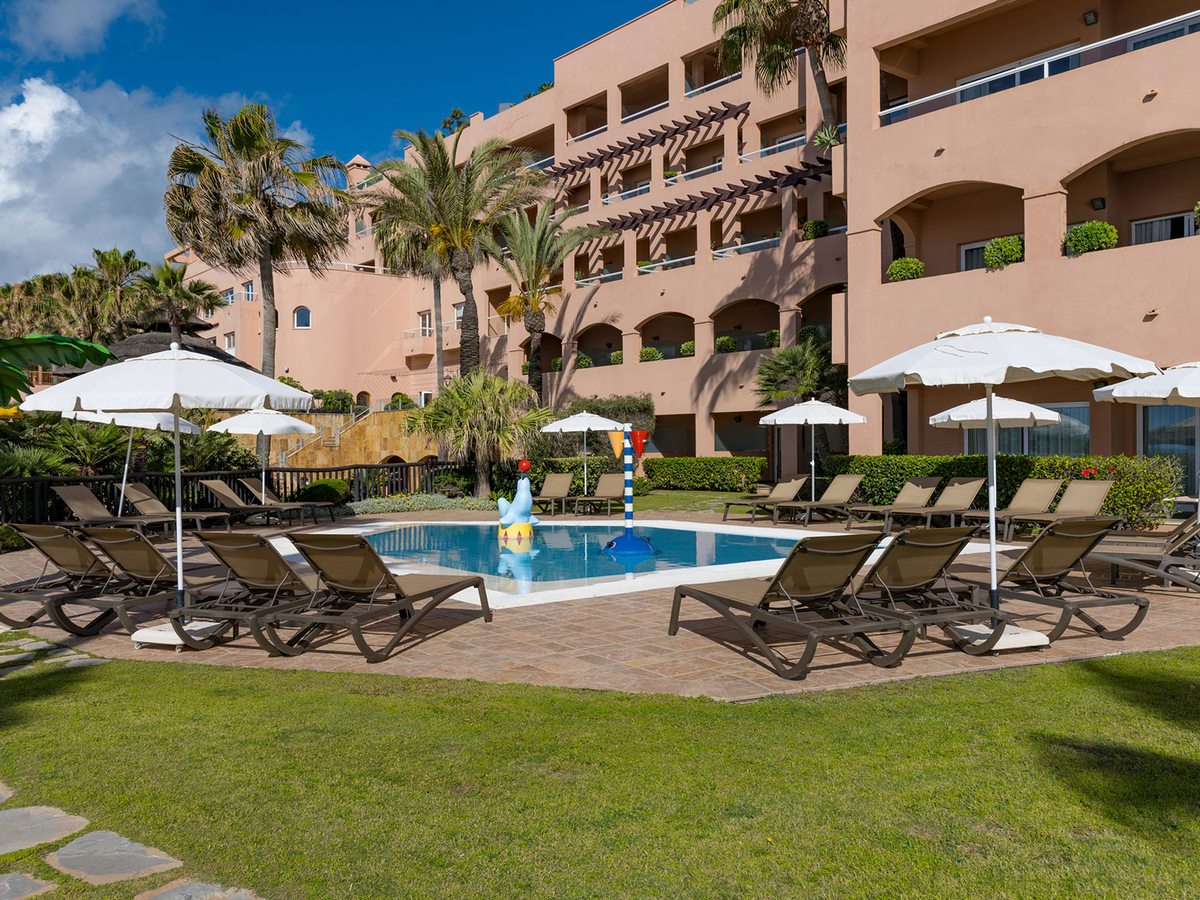A small shallow children's pool with mini water slides on a lawn beside the hotel building, with sun loungers and parasols.