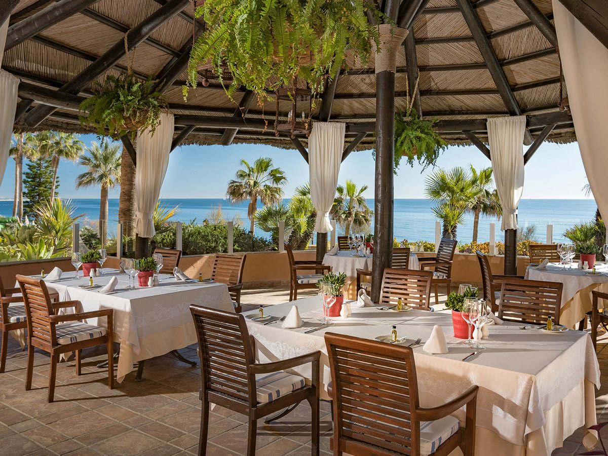 A covered outdoor restaurant terrace under a timber roof, with white-clothed tables and wooden chairs, hanging plants and palms with the sea beyond.