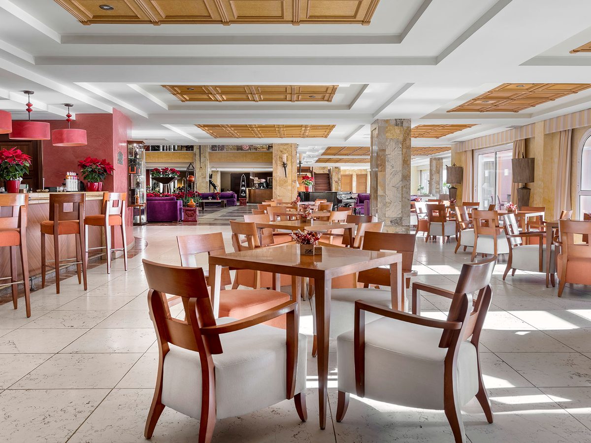 An indoor lounge bar with a counter, dining tables and wooden chairs under red pendant lights on a marble floor.