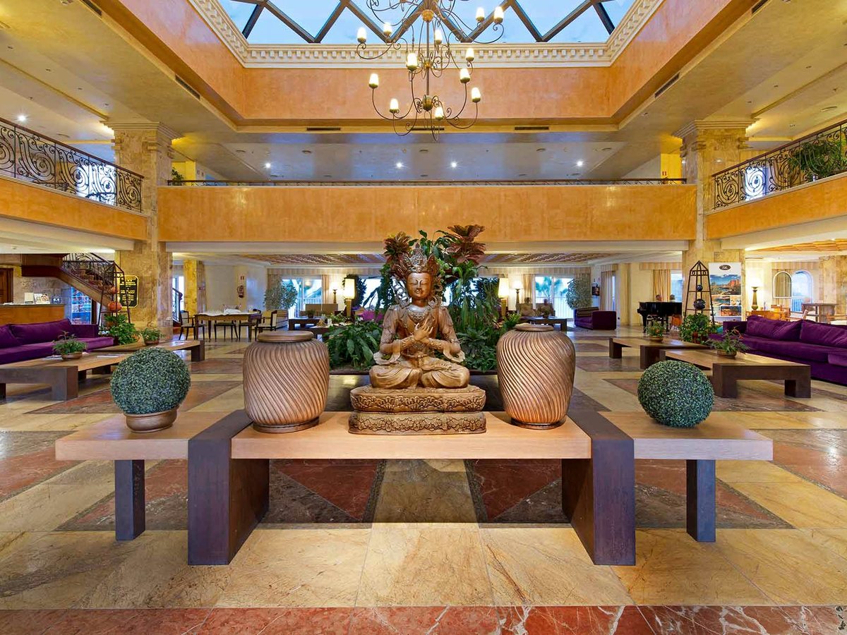 A grand hotel lobby atrium with a marble floor, a central sculpture, purple sofas, potted plants and balconied floors rising to a skylight.
