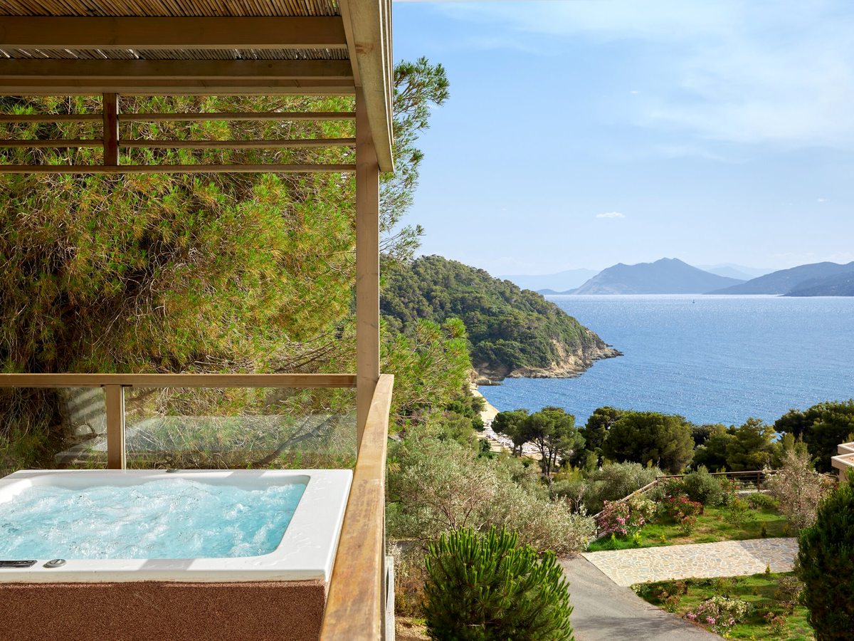 A sunken hot tub on a timber-decked terrace with a glass balustrade, looking out over pine trees to the sea and distant islands.