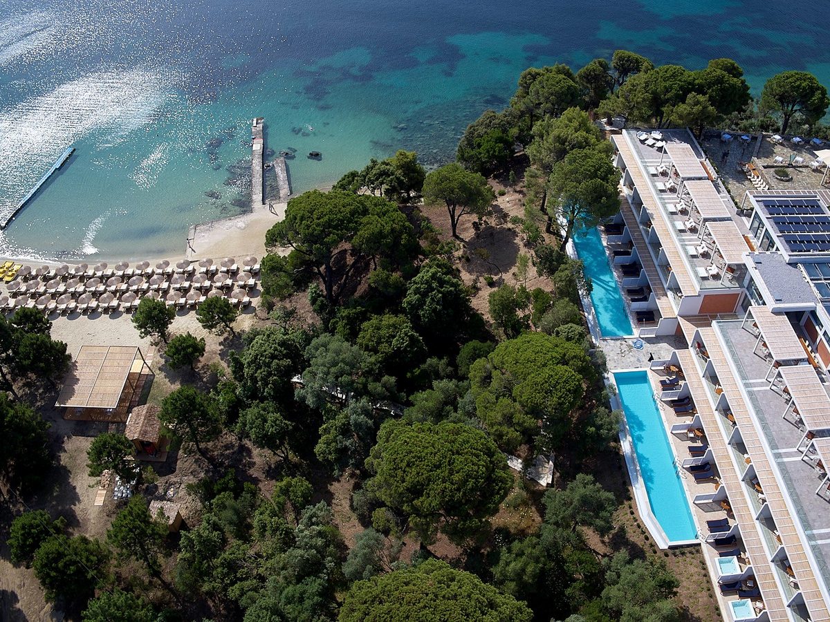 Aerial view of a sandy beach lined with straw parasols and a small stone jetty, beside the resort's beachfront building and its row of private plunge pools.