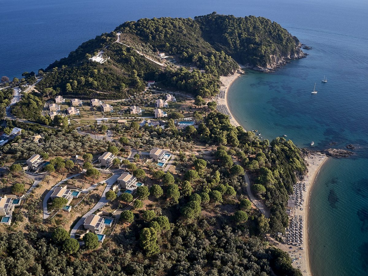 Aerial view of the whole wooded Punta peninsula at the south-western tip of Skiathos, ringed by calm blue sea with small islands offshore.