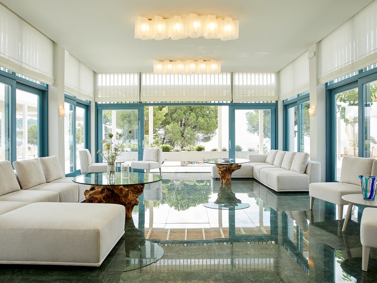 Bright open-plan lounge with white sofas and glass coffee tables on a green marble floor, with full-height blue-framed windows looking out to greenery and the sea.
