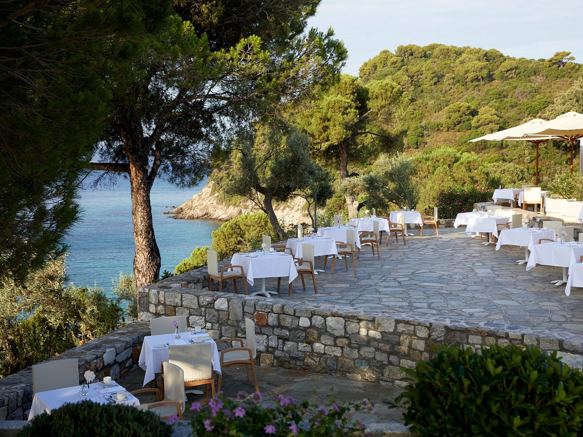 White-clothed dining tables on a stone terrace behind a low wall, shaded by a pine, above a small sandy cove and turquoise sea.
