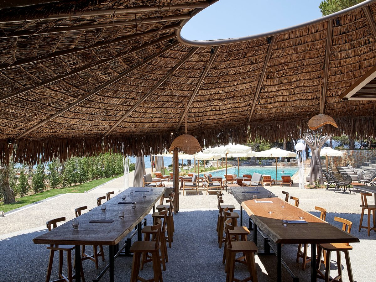 Long wooden tables and stools under a large thatched canopy, with parasols and a pool beyond among pine trees.