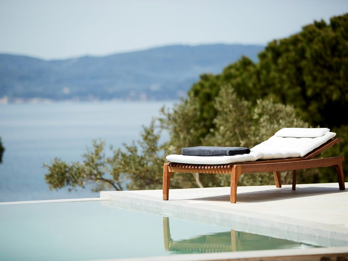 An infinity-edge pool with a single wooden sun lounger on the deck, looking across calm sea to distant hills.