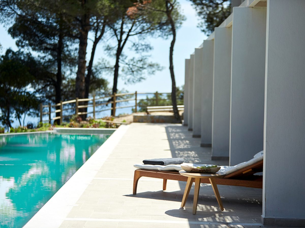 A long narrow pool running alongside a colonnade of white suite bays, with a sun lounger and low table on the paved surround under pines.