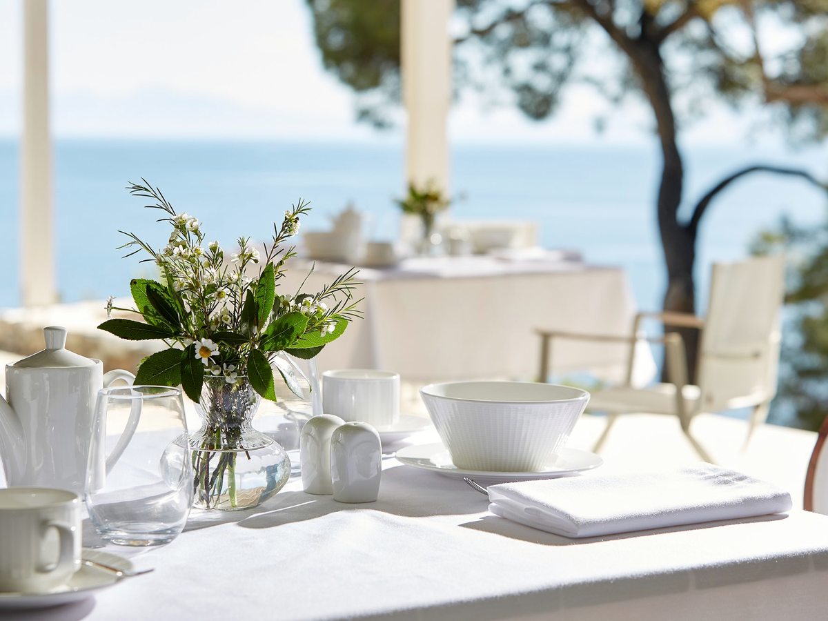 A breakfast table laid with white crockery, a coffee cup, a jug and a small vase of flowers, with the sea and trees beyond.