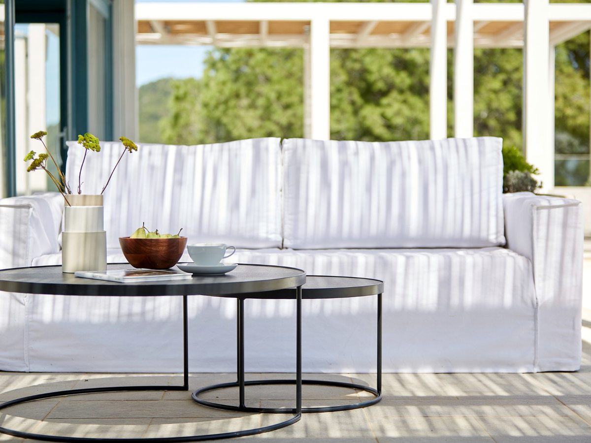 A covered outdoor lounge with a white cushioned sofa, round coffee tables and a fruit bowl, looking out through light curtains to trees and a hillside.