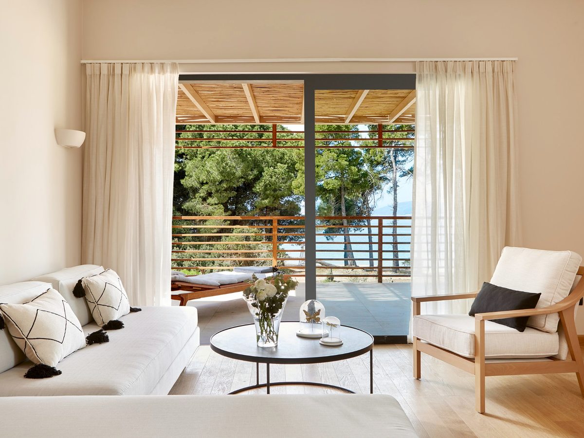 A suite sitting room with a pale sofa, armchairs and a low table, opening through glass doors to a balcony and the sea.