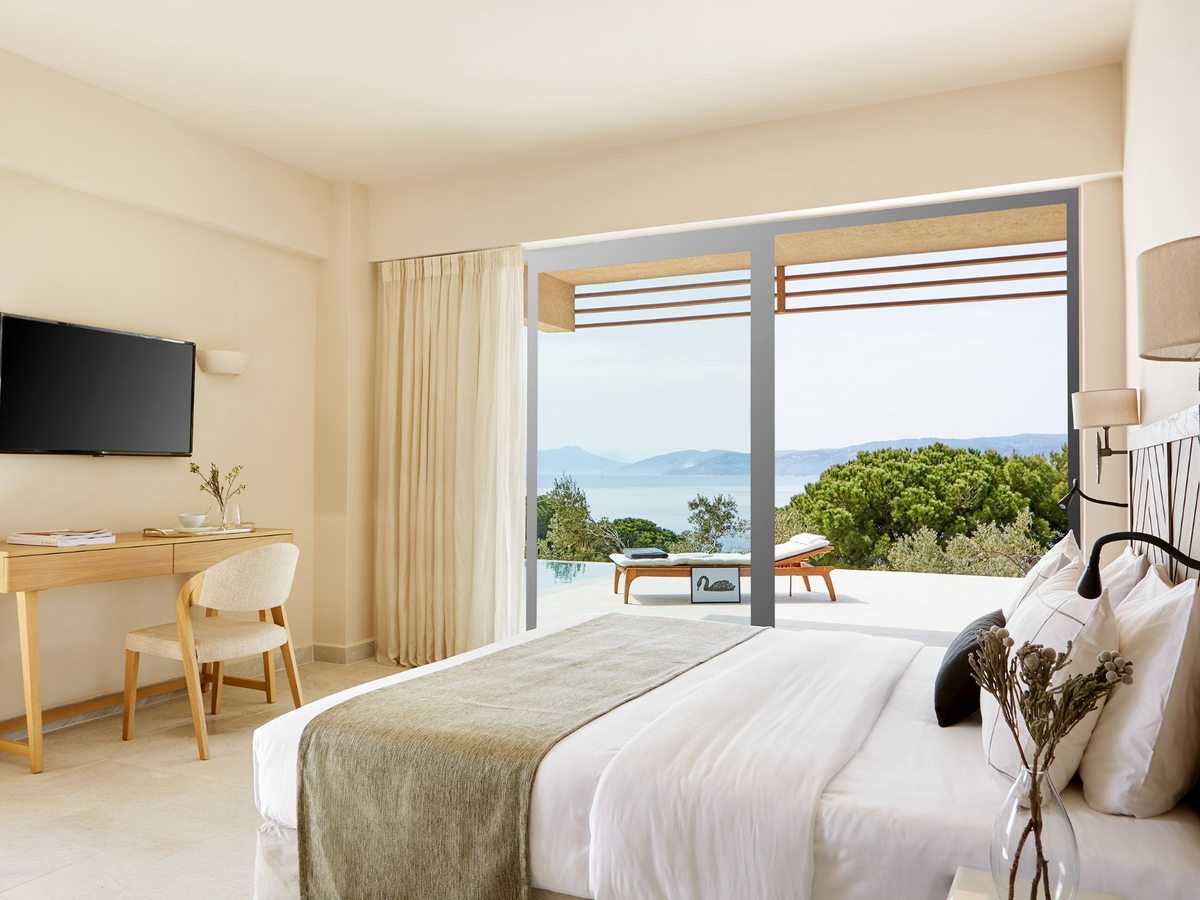 A bright bedroom with a bed, a writing desk and a wall-mounted television, glass doors opening to a balcony with sea and greenery beyond.