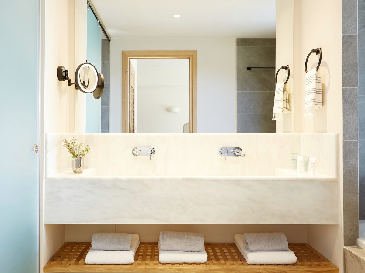 A long marble vanity with twin basins and a mirror, folded towels beneath and a small vase of greenery.