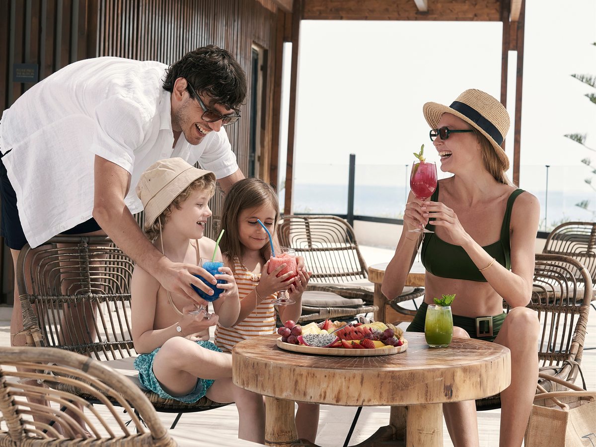 A family with drinks at a shaded terrace bar with wicker chairs around a round wooden table