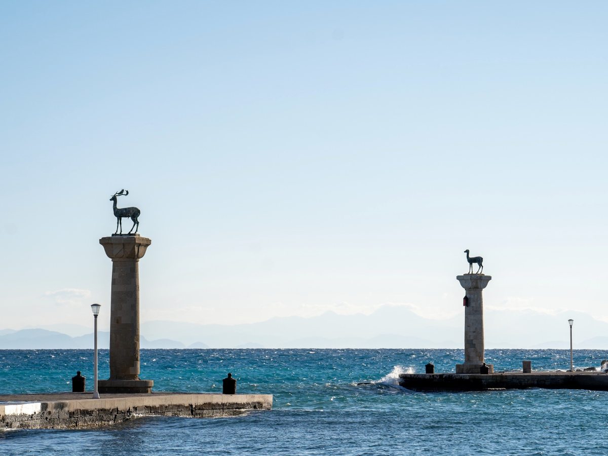 The bronze deer statues on stone columns at the entrance to Mandraki Harbour with a fort behind