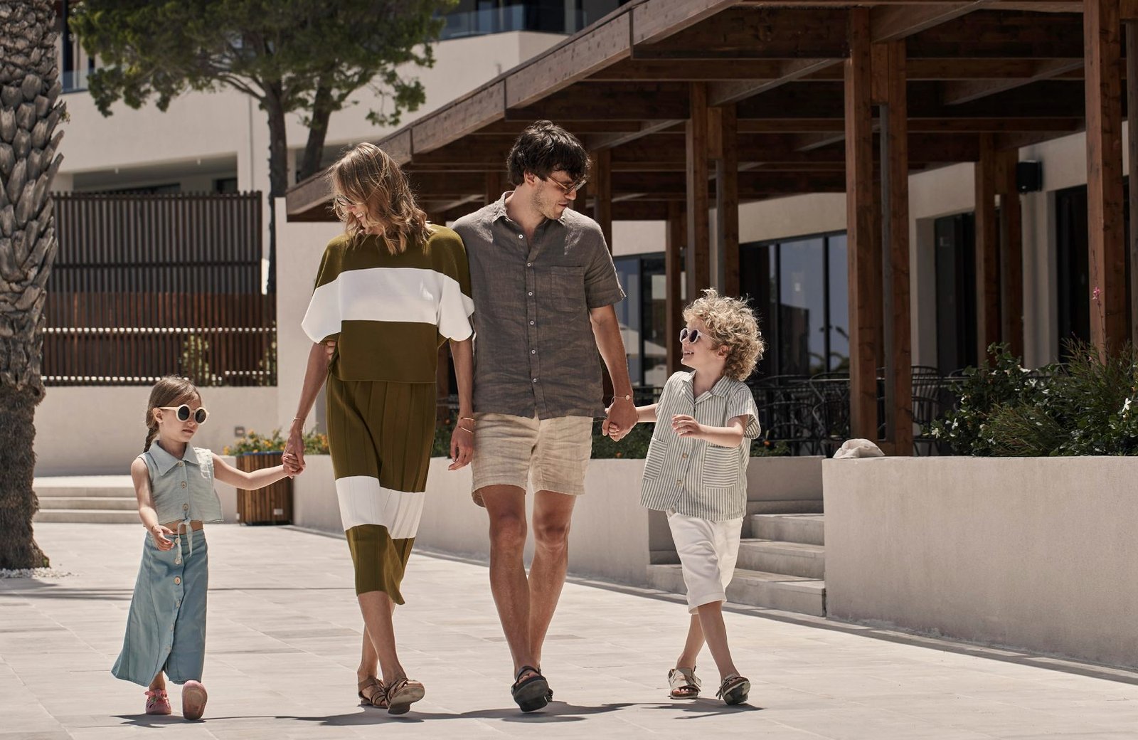A family of four walking hand in hand along a paved path in front of a low earth-toned resort building with a timber pergola at Ella Helea, Rhodes.