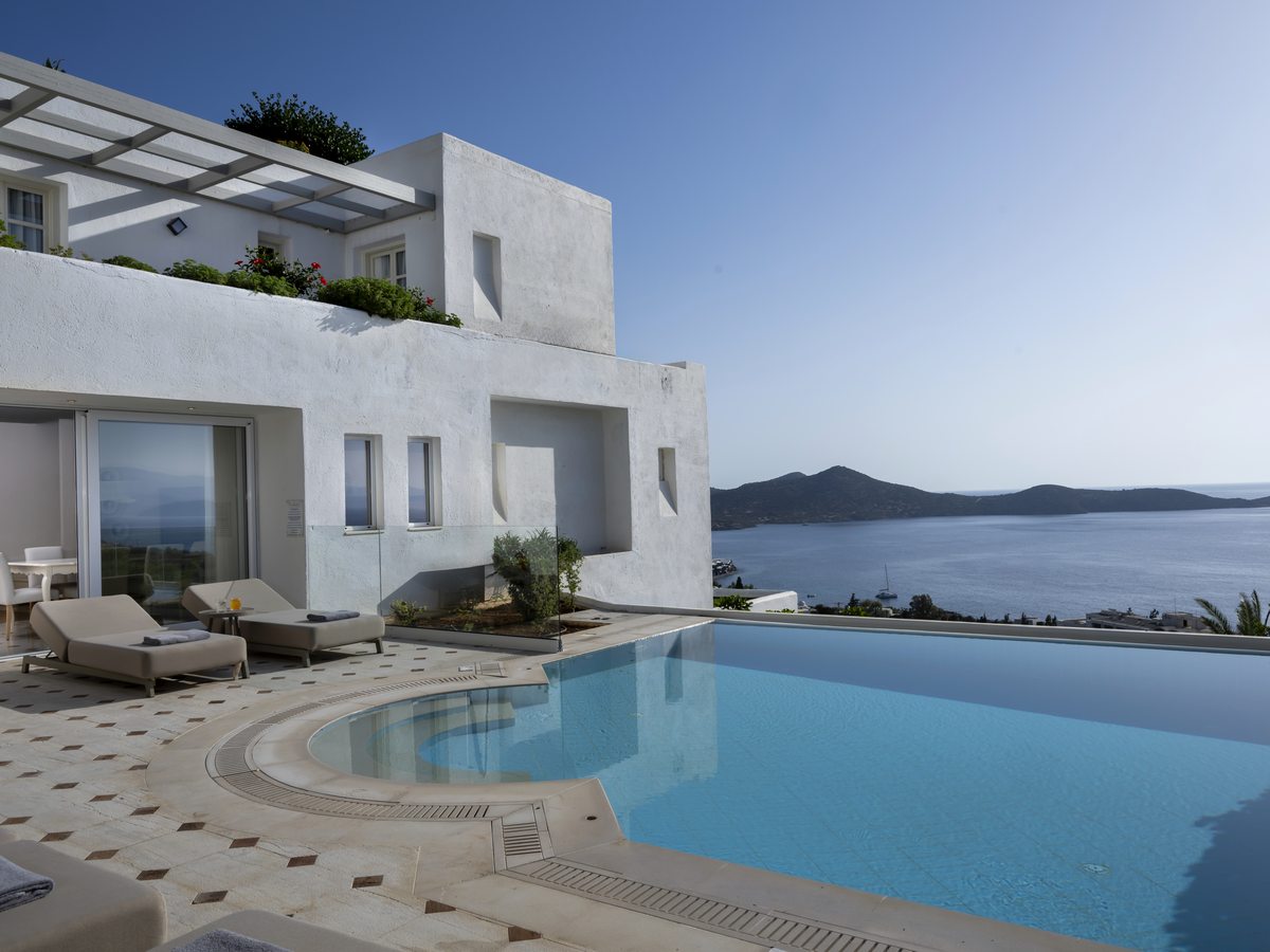 White villa with an infinity pool and sun loungers on a tiled terrace, looking out over the Gulf of Mirabello to distant hills.