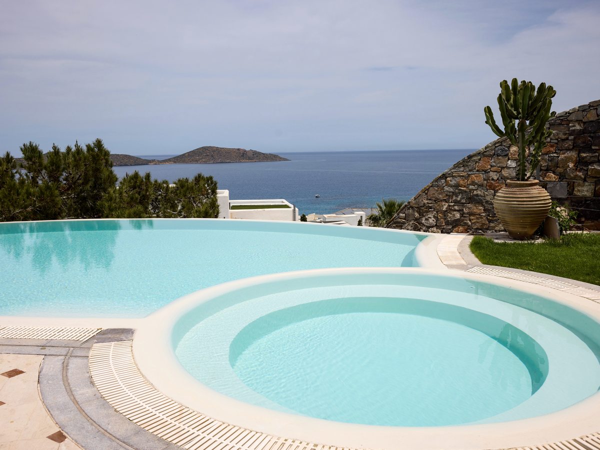 Curved infinity pool with a circular Jacuzzi section in the foreground, a stone wall and planted urn to one side and the sea and an island beyond.