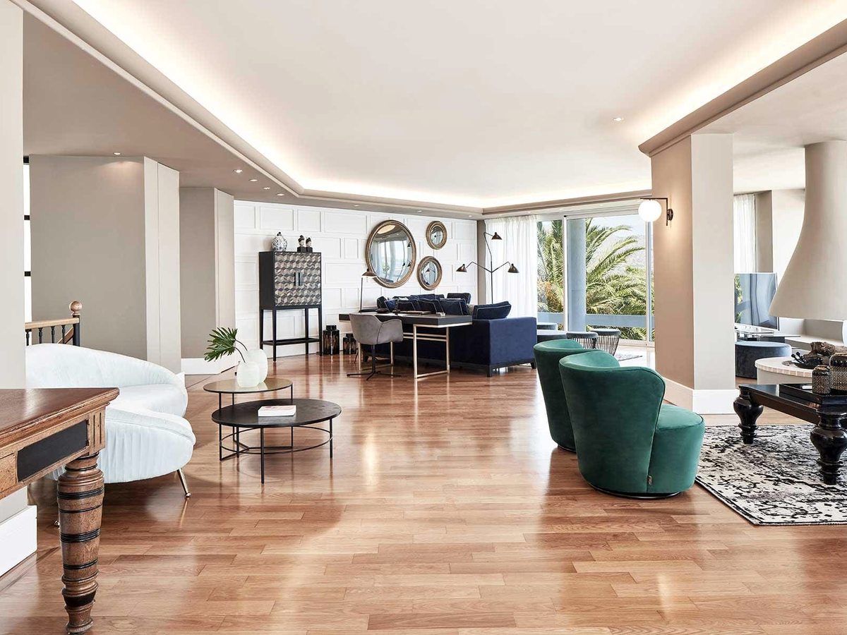 Large open-plan living room with a wood floor, a navy sofa, green velvet armchairs, antique-style console and mirrors, and glass doors to a garden terrace.