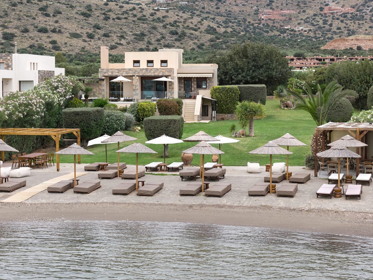 Row of thatched umbrellas and cushioned sun loungers on a narrow sand-and-shingle beach in front of a lawn and a stone tavern building, hills behind.
