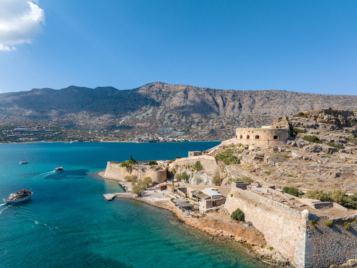 Aerial view of Spinalonga island with its Venetian fortress walls and ruined stone buildings, small boats on the turquoise water and the mainland behind.