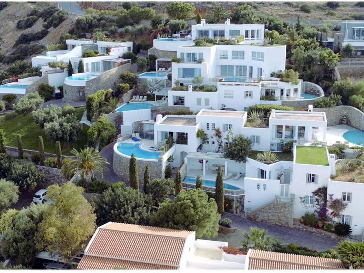Aerial view of white flat-roofed villas stepping down a steep hillside, each with a small private pool, among olive trees and dry scrub.