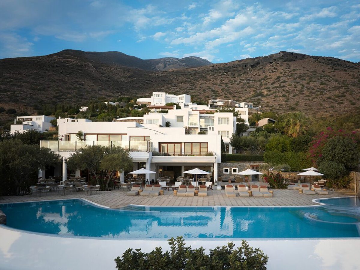 White multi-level hotel building on the hillside above a large curved pool ringed with sun loungers and parasols, bare mountains behind.