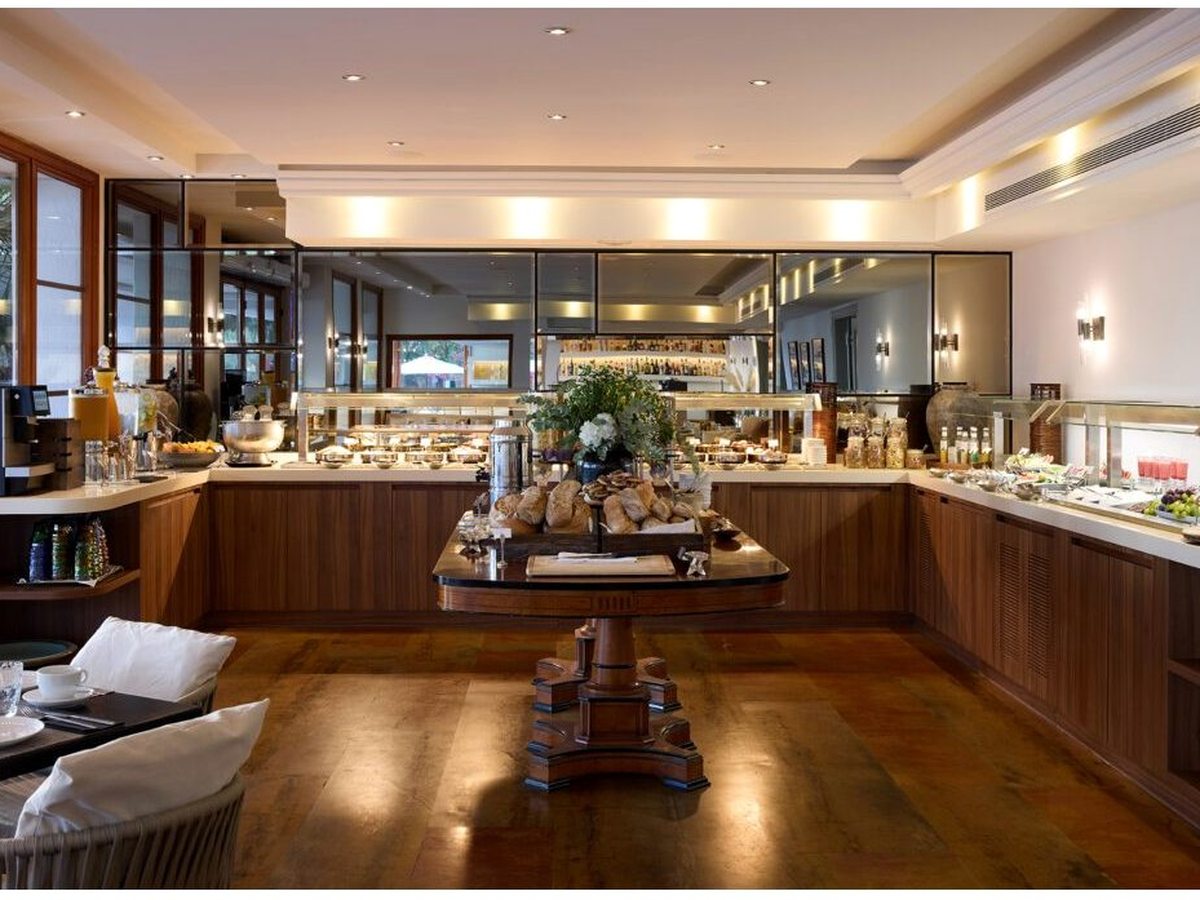 Warmly lit buffet room with a central wooden table of breads and pastries, a coffee machine and counters of fruit and juices under mirrored cabinets.
